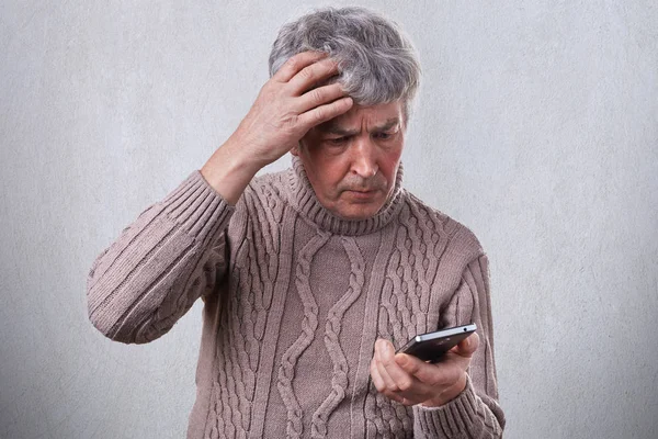 A troubled mature man holding his hand on his gray head while using smartphone reading some news in the Internet. A man having shock while looking into his smartphone. — Stock Photo, Image