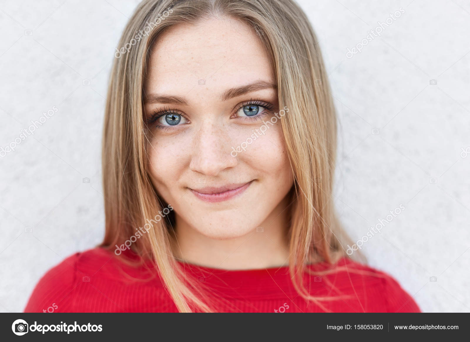 Close up of blonde woman with blue eyes, long eyelashes and dimples looking  pleasantly into camera rejoicing her life. Pretty young fair-haired female  in red clothes posing against white concrete wall Stock