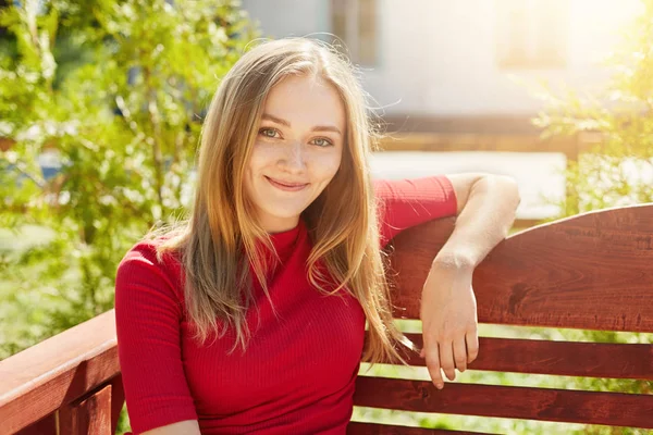 Retrato de mulher tranquila despreocupada com o cabelo claro tendo descanso no parque enquanto sentado no banco de madeira desfrutando de bela natureza e ar fresco. Menina atraente descansando ao ar livre durante suas férias de verão — Fotografia de Stock