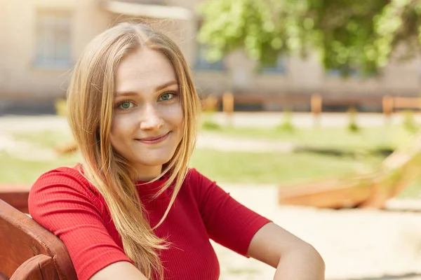 Νεαρή μαθήτρια με ξανθά μαλλιά και σαγηνευτική εμφάνιση στο κόκκινο φόρεμα κομψό κάθεται σε εξωτερικούς χώρους σε ξύλινο πάγκο περιμένοντας κάποιον θέτοντας φόντο παιδική χαρά έχοντας θετική έκφραση — Φωτογραφία Αρχείου