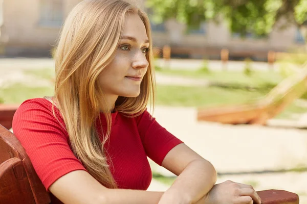 Portrait latéral d'une jeune femme coûteuse aux cheveux blonds portant un pull rouge assis à l'extérieur sur un banc confortable en bois ayant un regard profond et réfléchi en pensant à quelque chose d'important — Photo
