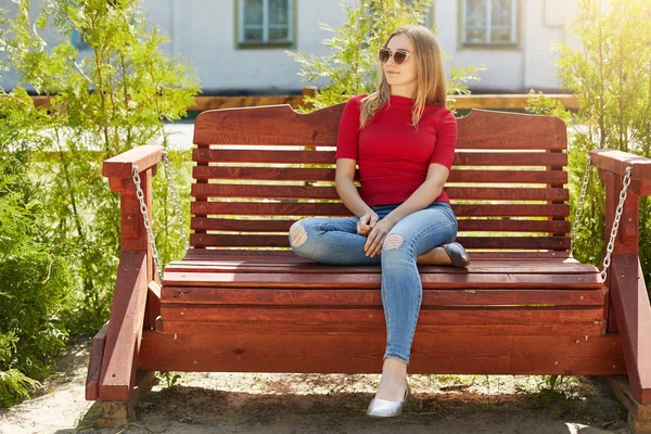 Открытый портрет беззаботной стильной девушки-подростка в красном свитере, джинсах и солнцезащитных очках, сидящей на большой удобной скамейке и глядящей в сторону в ожидании своего бойфренда. Человек, стиль и молодость — стоковое фото