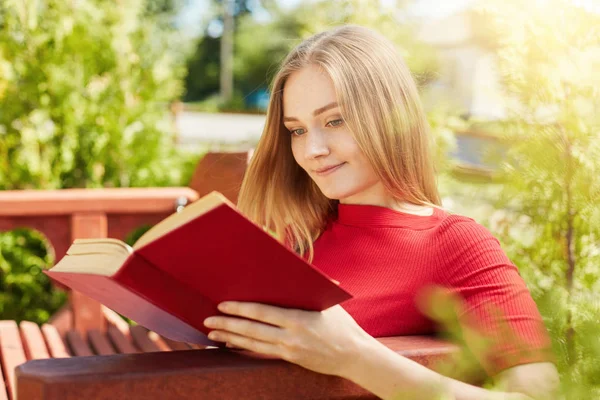 Outdoor portrait of young teenage girl with fair hair dressed in red blouse sitting at wooden bench and reading her favourite book admiring summer weather and sunshine. Relaxation and rest concept — Stock Photo, Image