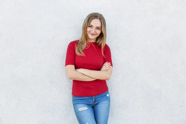 Όμορφη γυναίκα σε κόκκινο πουλόβερ και τζιν στέκεται σταυρωμένα χέρια λευκό τοίχο που κοιτάζουν κατευθείαν την κάμερα έχοντας ευτυχισμένη έκφραση. Ξανθούς γυναίκα που φοράει μοντέρνα ρούχα στέκεται ενάντια στο λευκό — Φωτογραφία Αρχείου