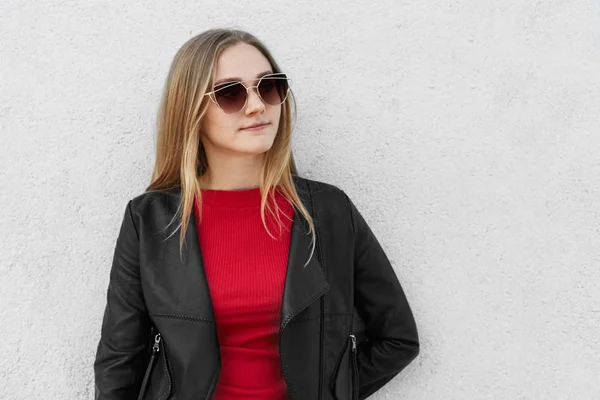 Sexy mujer rubia en gafas de sol y abrigo de cuero negro de pie contra la pared de hormigón blanco con espacio en blanco para su anuncio mirando a un lado con expresión reflexiva y aspecto agradable — Foto de Stock