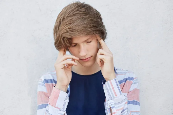 Pensive concentrated teenage boy with stylish haircut looking down thinking over something important. Fashionable male holding fingers on temples trying to concentrate having very serious look — Stock Photo, Image