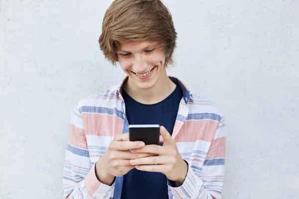 Adolescente alegre con peinado de moda sosteniendo mensajes de escritura de teléfonos inteligentes modernos o jugando juegos en línea sonriendo agradablemente teniendo hoyuelos en su cara de pie sobre fondo de hormigón blanco — Foto de Stock