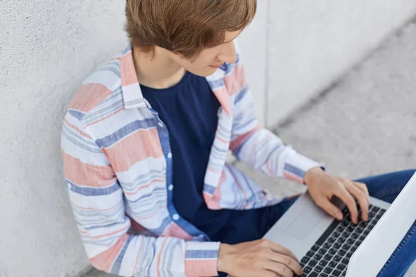 Обрезанный снимок стильного студента-мужчины, работающего на курсовой бумаге, печатающего на ноутбуке необходимую информацию. Мальчик, использующий компьютер для серфинга в социальных сетях с бесплатным подключением к Интернету — стоковое фото