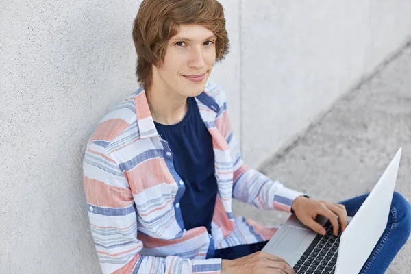 Fashionable serious teenager sitting on floor holding laptop typing something on keyboard looking in camera. Male college student having rest watching video or reading news online. Technology concept — Stock Photo, Image