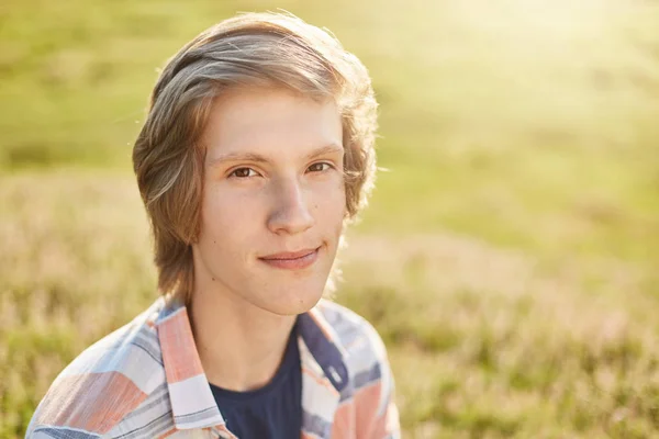 Close up portrait of attractive teenager with dark eyes, pure skin and trendy hairstyle wearing shirt looking directly into camera while sitting at greenland breathing fresh air and admiring sunlight — Stock Photo, Image