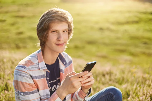 Pleased young boy with dark narrow eyes and stylish hair sitting at meadow holding cell phone checking his e-mail or reading news using free internet connection while resting at beautiful green field — Stock Photo, Image