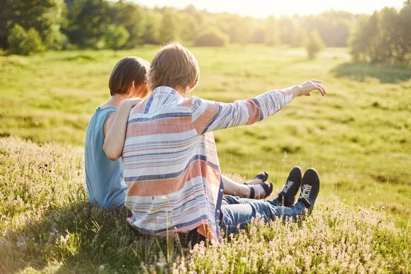 Ragazzo adolescente mostrando qualcosa con la mano alla sua ragazza che indica in lontananza mentre seduto al campo verde. Fidanzata romantica e fidanzato che si abbraccia mentre si gode le vacanze estive all'aperto — Foto Stock