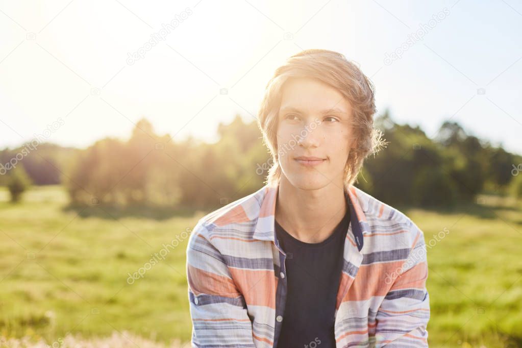 Dreamy attractive boy with light hair, dark eyes and thin lips wearing shirt sitting over green nature background looking aside with face full of dreams or thoughts relaxing after hard day at nature