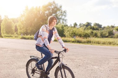 Son moda saç modeli ile yakışıklı giyen gömlek ve sırt çantası olan kot binicilik bisiklet sakin atmosferi mesafe içine bakarak zevk yol geri. Genç çocuk muhteşem kırsal kesimde Bisiklete binme