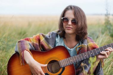 Ciddi erkek çekici görünüme çayır şarkıyı çalmak için akustik gitar ile oturan güneş gözlüğü takıyor. Yeşil çimlere otururken sakin atmosferi tadını genç kadın gitarist