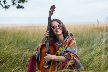 Yetenekli genç erkek rahat geniş açık havada dinlenmek için ve frsh hava, muhteşem manzara keyfi sahip kameraya gülümseyen gitar tutan giyinmiş. Yaşam tarzı ve dinlenme kavramı