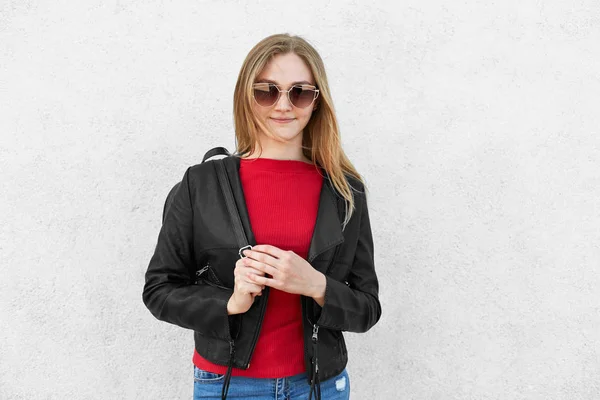 Retrato horizontal de una mujer de moda con chaqueta negra, suéter rojo y gafas de sol de moda con mochila en la espalda aislada sobre una pared de hormigón blanco. Adolescente chica en ropa moderna — Foto de Stock