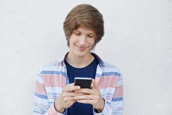 Hipster chico sosteniendo el teléfono inteligente escribiendo mensajes a su novia que ha complacido la expresión de decirle acerca de su amor. Adolescente alegre usando un dispositivo moderno mientras está de pie sobre una pared de hormigón blanco — Foto de Stock