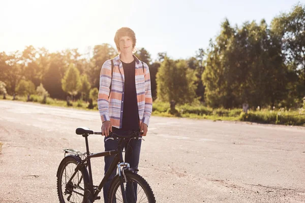 Şık saç gömlek ve güzel doğa hayran gezi sahip bisiklet ile ayakta kot giyen genç çocuk. Geri kalan uzun yolculuk sonra sahip yolda duran yakışıklı bisikletçi. Spor, eğlence