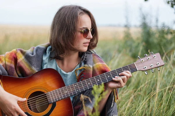 Belle femelle avec une coiffure courte portant des lunettes de soleil élégantes reposant à l'extérieur jouant de la guitare en profitant de ses vacances d'été. Musicienne dans des lunettes de soleil élégantes jouant de l'instrument de musique — Photo