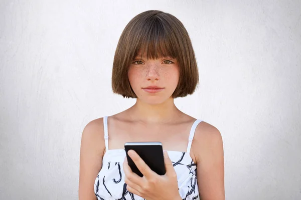 Dzieciństwo w epoce cyfrowej. Ładna dziewczyna z krótką fryzurę stylowe, ciemne głęboko osadzone oczy i piegi na sobie ładny strój, trzymając smartfon w jej ręce, grając w gry online na białym tle na białej ścianie — Zdjęcie stockowe