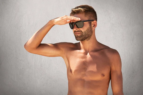 Retrato de homem bonito sem barba nu em óculos de sol, mantendo a mão na testa, olhando para a distância posando contra a parede de concreto cinza. Esportista forte que tem férias de verão vai nadar — Fotografia de Stock