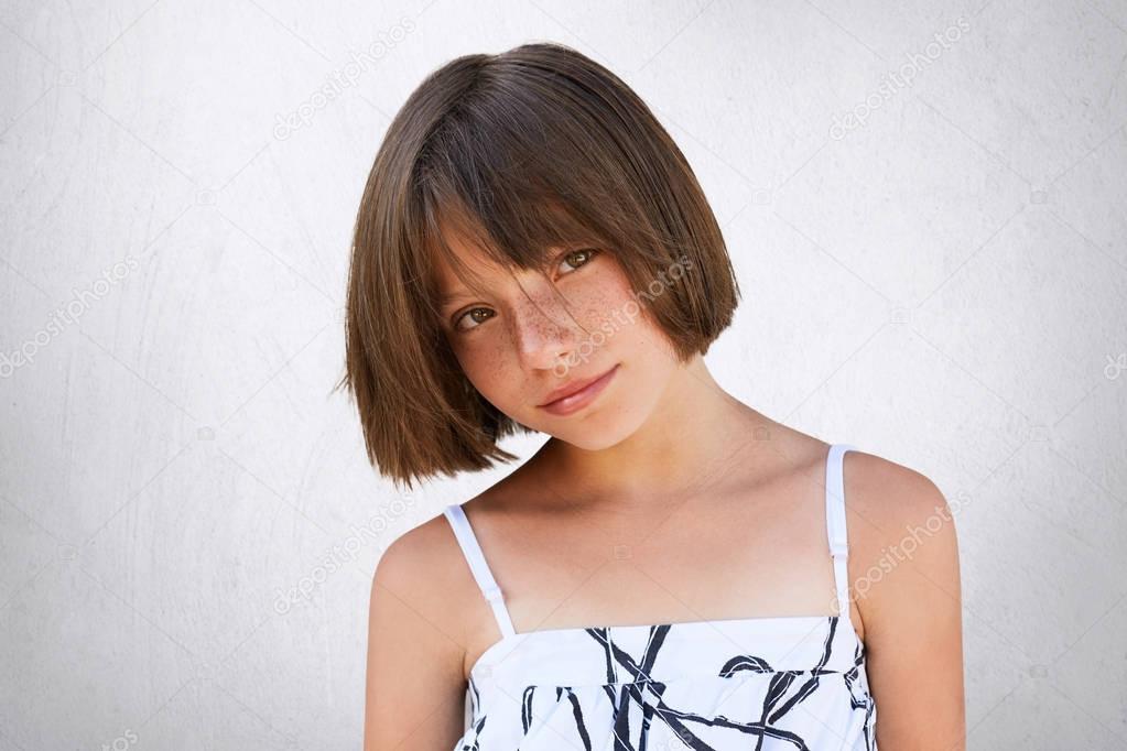 Pretty little girl with freckles, dark eyes and short hairstyle wearing summer white dress posing against white background having confident and serious look. Children, lifestyle, beauty concept