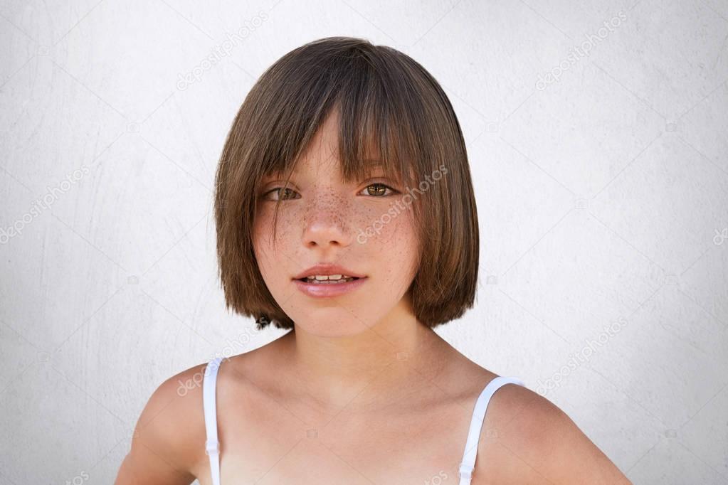 Adorable little child with brown charming eyes, freckled skin and thin lips having stylish hairdo, wearing summer clothes, looking directly into camera while posing against white background.