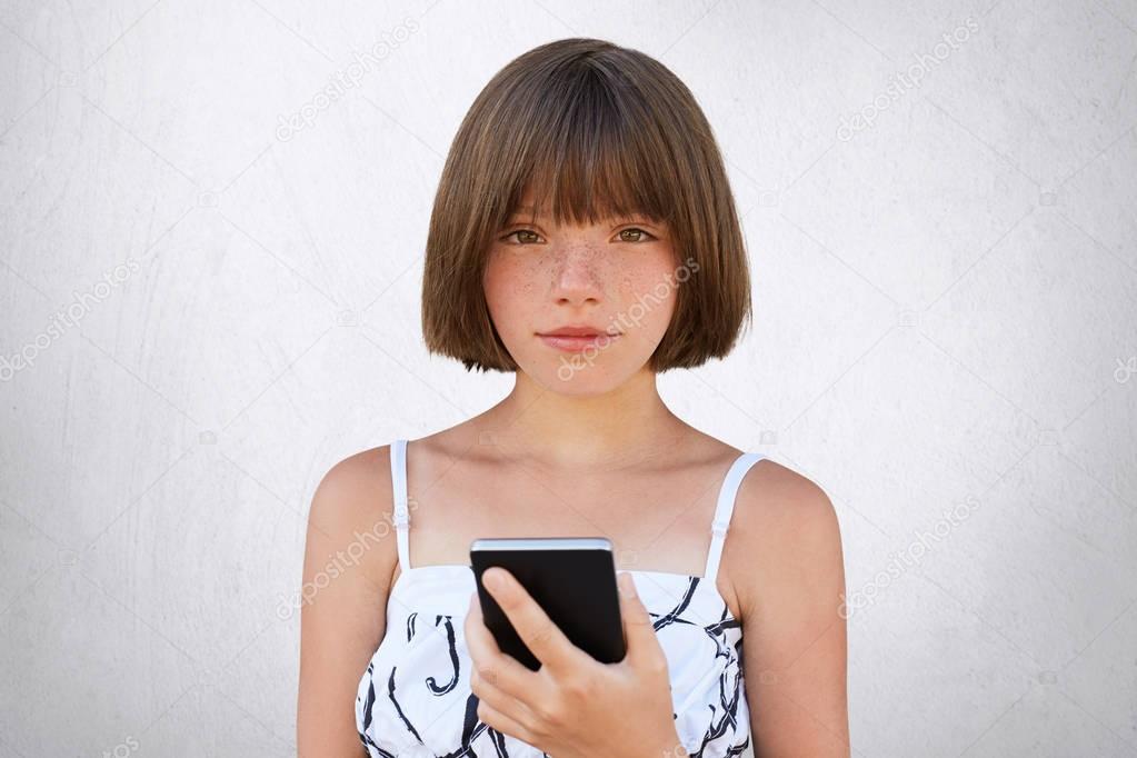 Childhood in digital age. Cute girl with short stylish hairdo, dark deep-set eyes and freckles wearing nice dress, holding smart phone in her hands, playing games online isolated on white wall