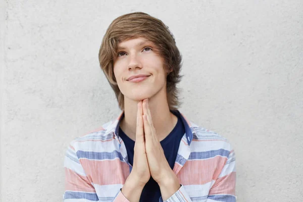 Jonge Europese tiener met trendy kapsel houden zijn hand samen opzoeken met trouwe look, bidden voor goed geluk op examen. Jongen met ogen vol geloof geïsoleerd over grijze achtergrond — Stockfoto