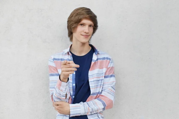 Menino adolescente confiante com penteado elegante vestindo camisa casual apontando com o dedo para a câmera enquanto mostra algo. Garoto elegante com aparência atraente posando contra a parede de concreto cinza — Fotografia de Stock