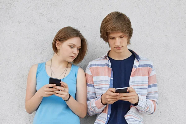 Two teenagers using modern gadgets, playing games online while standing against grey concrete wall. Pretty girl in blue dress looking in cell phone of her fashionable boyfriend with curious look — Stock Photo, Image
