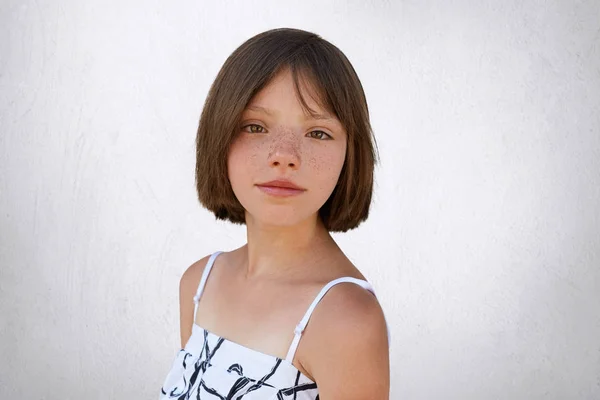 Good-looking brunette kid with freckles and short hair posing against white concrete wall dressed in white dress. Little child with dark wide eyes and freckled skin isolated over white background — Stock Photo, Image