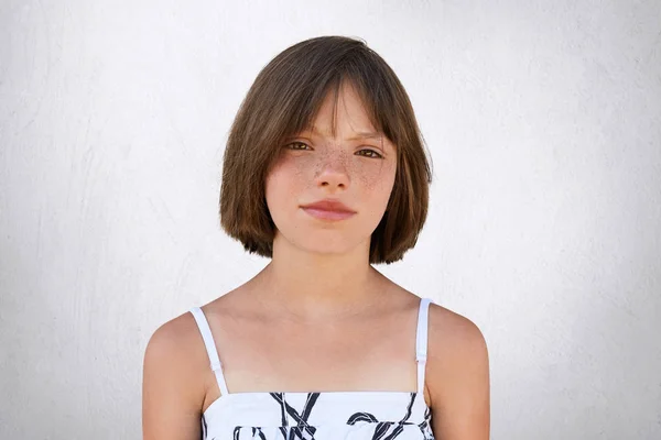 Freckled girl with hazel eyes and dark short hair, looking with displeasure into camera while posing against whiye concrete wall. Beautiful little girl with stylish hairdo wearing summer dress — Stock Photo, Image