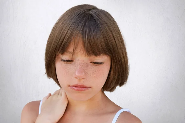 Close up of attractive little child with freckles and dark short hair keeping her hand on neck, looking seriously down, having thoughtful expression while posing against white concerete wall — Stock Photo, Image