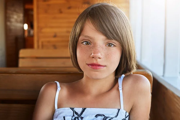 Children, rest, relaxation concept. Pleasant-looking freckled girl with short dark hair, wearing summer dress while sitting at deck on wooden bench, looking directly into camera with her shining eyes — Stock Photo, Image