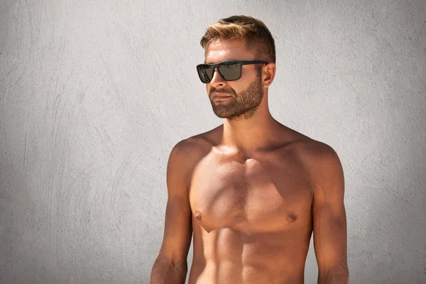 Portrait taille haute d'un homme macho attrayant avec des poils dans des lunettes à la mode, debout nu contre un mur blanc gris, regardant de côté avec une expression confiante, démontrant son corps bronzé et sportif — Photo