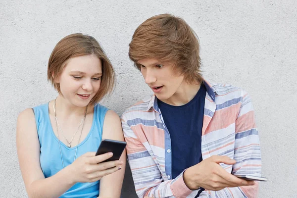 Teenage girl with trendy hairstyle wearing blue dress, holding her smart phone, showing fotos to her male friend, who is looking attentively. Teenagers using modern devices. People, modern technology — Stock Photo, Image