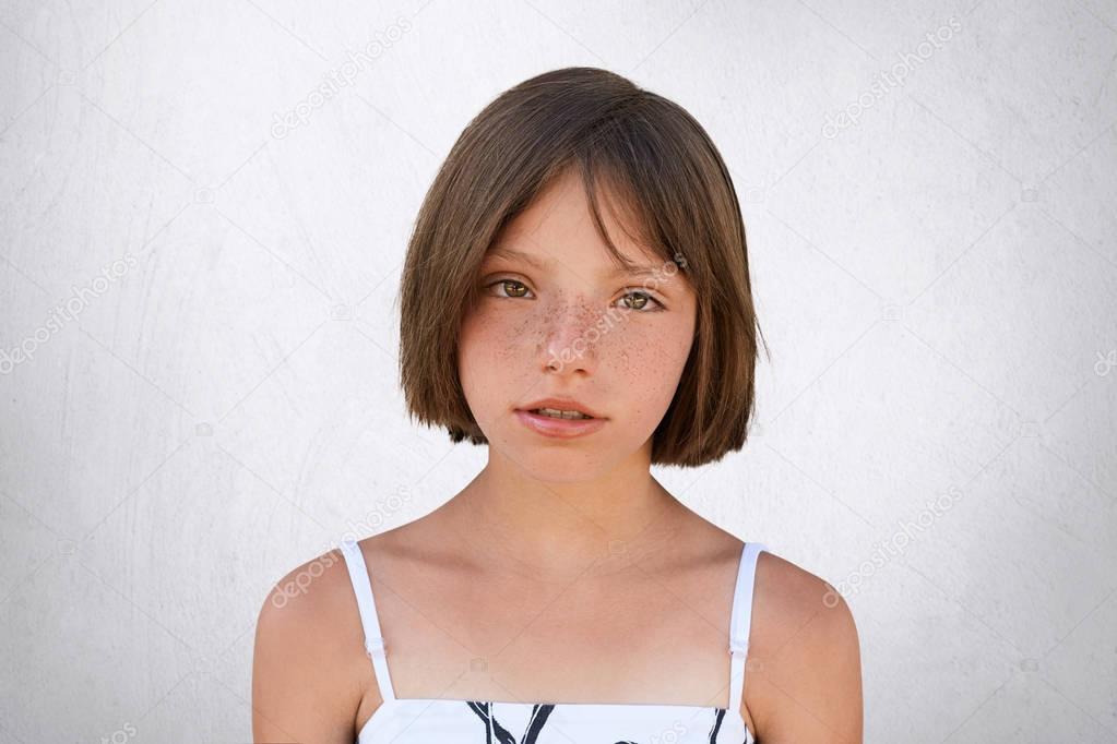Serious freckled girl with bobbed hair and dark eyes looking directly into camera, isolated over white background. Stylish adorable little girl in white dress. People, childhood, emotions concept