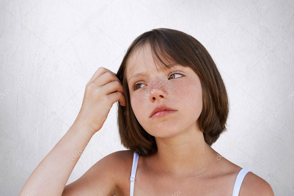 Serious little freckled child, keeping her hand on hair, havin thoughtful expression, looking aside. Beautiful girl posing against white background. Beauty, childhood, facial expression concept
