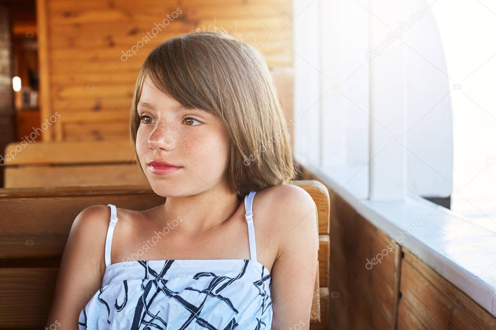 Pensive little child with bobbed hair resting on wooden deck, looking aside while dreaming about something. Pretty girl in white and black dress sitting at wooden bench and table on big ship