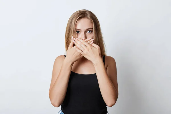 Blonde vrouw met glanzende ogen, die betrekking hebben op haar mond met handen terwijl het proberen om het geheim van haar beste vriend houden van stilte en niet vertellen. Jonge vrouwelijke model maken hush teken, isoated over witte muur — Stockfoto