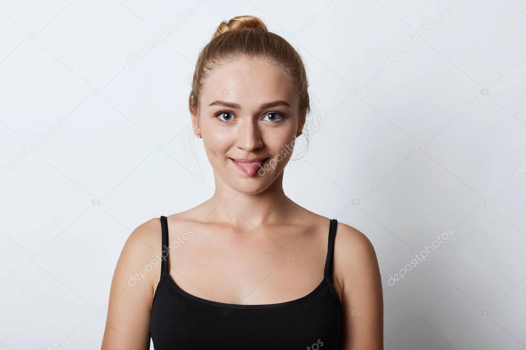Indoor shot of funny blonde female with hair knot, showing tongue while arguing with someone, trying to persuade that she is right. Beautiful female model showing tongue into camera isolated on white