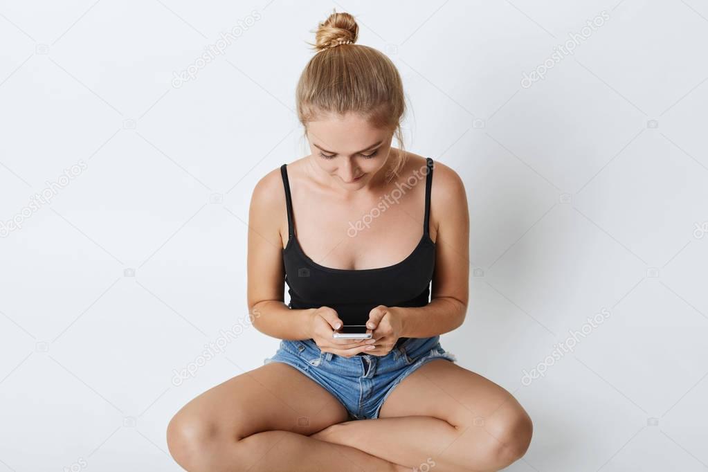 Carefree female with hair bun wearing casual t-shirt and denim shorts, relaxing on floor, typing messages while using mobile phone, communicating with her best friend using free internet connection