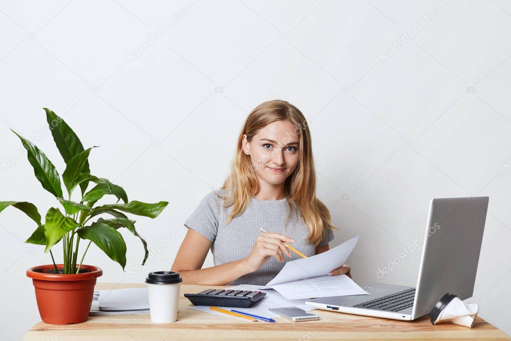 Young blonde businesswoman sitting at her workplace while making business report, calculating annual figures, reading documents and using modern technologies for her work, drinking takeaway coffee