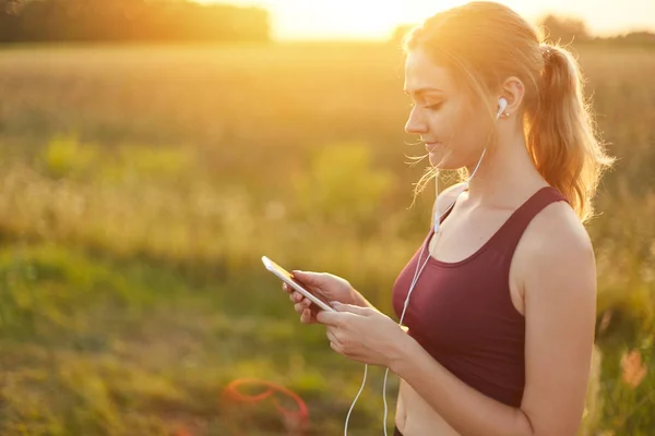 Pelari wanita menikmati suasana tenang saat jogging di lapangan, mendengarkan lagu favoritnya di earphone. Perempuan muda masuk untuk olahraga. Orang, gaya hidup aktif, konsep kesehatan dan kebugaran — Stok Foto