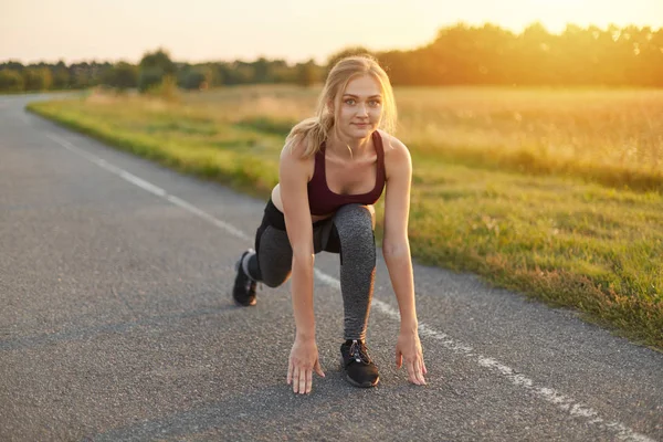 Horizontale portret van blonde vrouw, dragen van de top, broek en sport schoenen, doen van oefeningen op weg, haar benen strekken, zijnde vol energie. Sportieve vrouw warming-up haar benen voordat u — Stockfoto