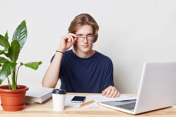 Portrait of convinced self assured male enterpreneur looks seriously through glasses, surrounded with modern laptop, mobile phone and documents, waits for business partner. Career and job concept