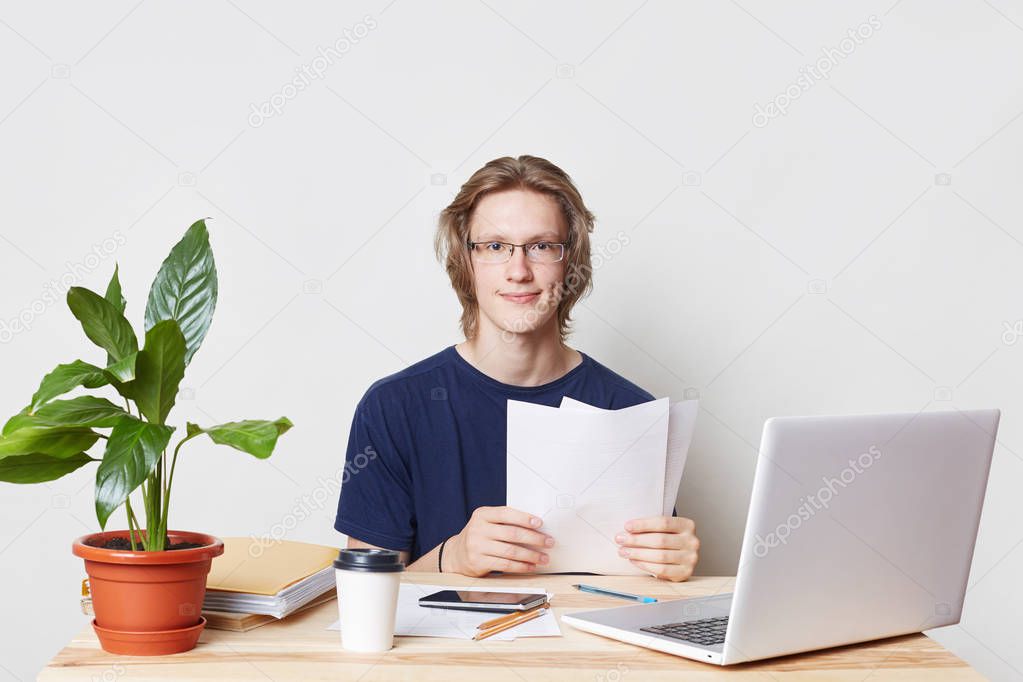 Hard working professional business worker sits at work place, reviews his accounts, studies documents, looks directly into camera with delightful expression, uses modern technologies for work