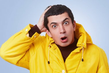 Surprsied desperated yakışıklı adam dinleme cihazı gözler ve yaygın olarak açılan ağız ile görünüyor, kukuleta ile sarı anorak giyer, uyuşukluk, mavi stüdyo arka plan üzerinde izole olmak anlamıyor musun,